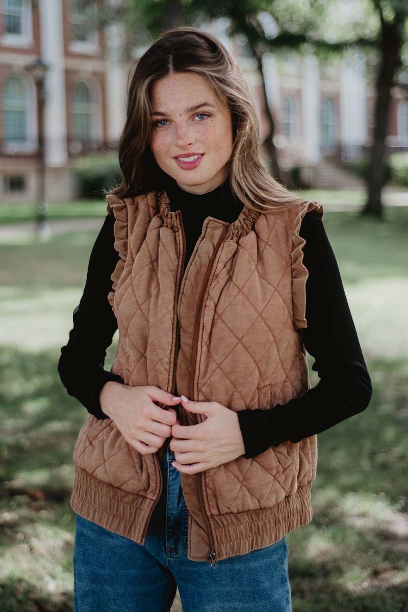 A person stands outdoors in a park-like setting, wearing a brown quilted vest over a black top.