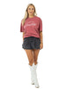 A woman with long blonde hair smiles as she faces forward, wearing the Katydid Game Day Social Club Graphic Tee in pink, a dark pleated mini skirt, and white cowboy boots against a white background.