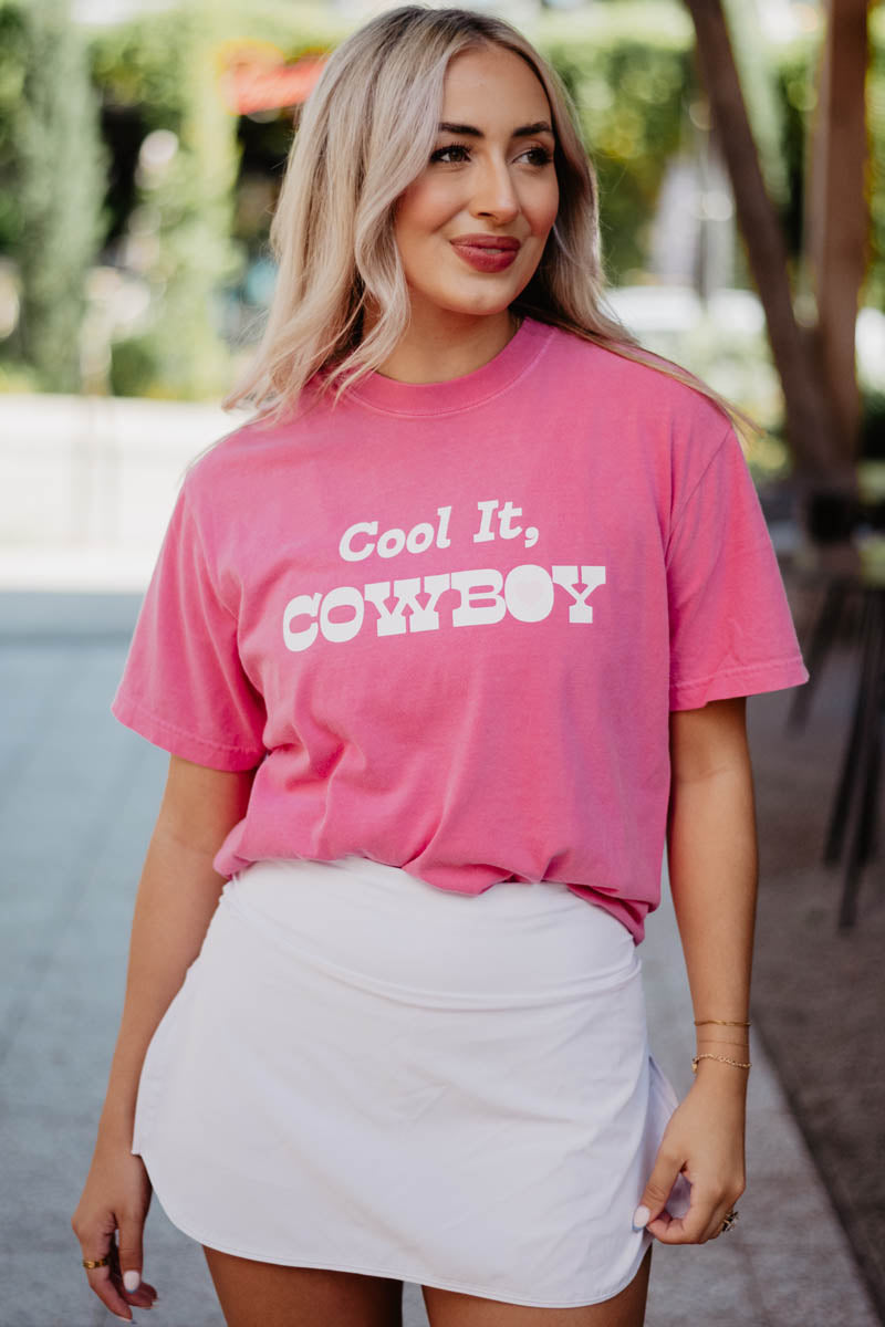 A woman with long blonde hair stands outdoors in a Katydid Cool it, Cowboy graphic t-shirt and a white skirt, smiling slightly. The sunlit background is blurred with greenery.