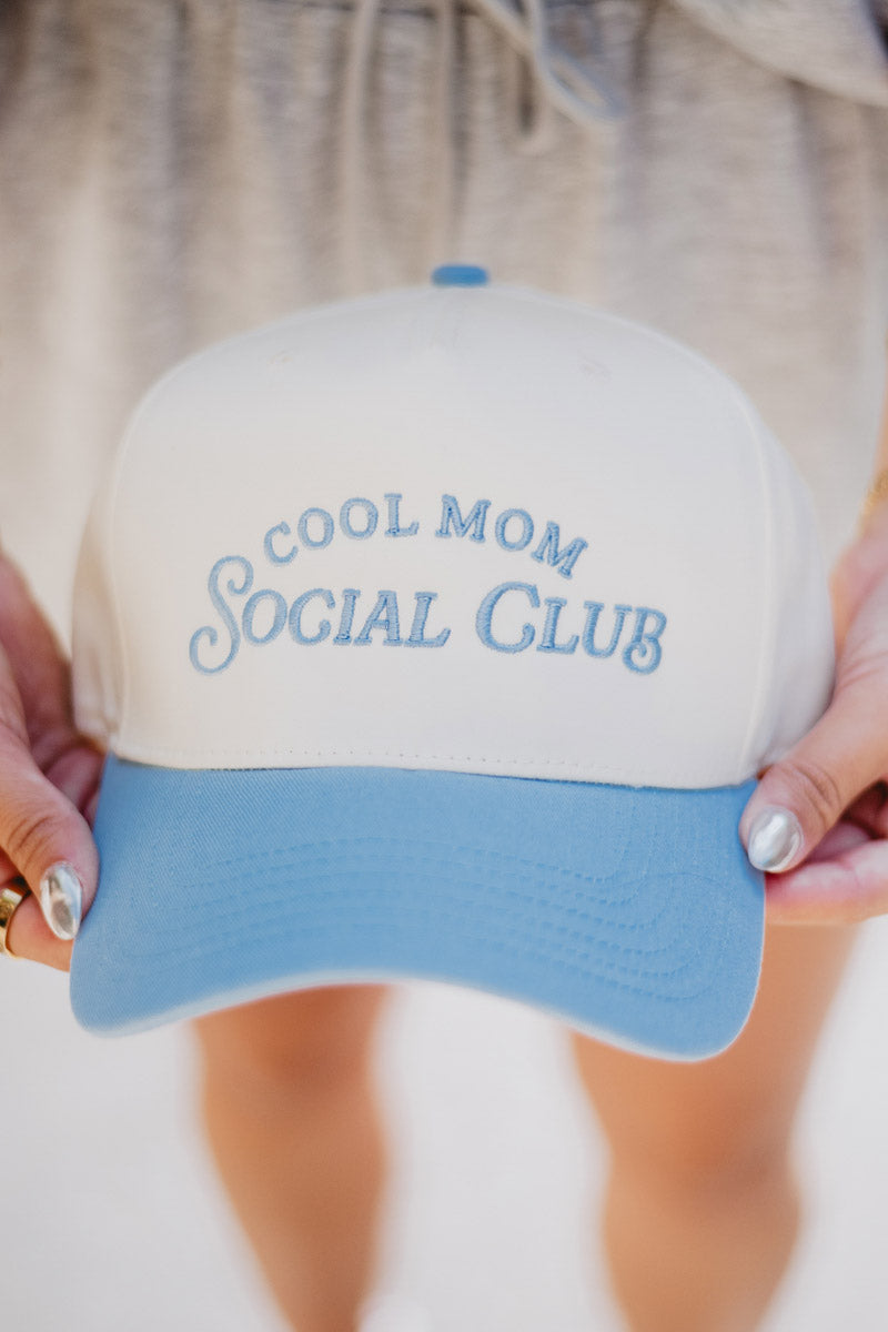 Close-up of a white and blue hat with "Cool Mom Social Club" in light blue text. The hat is held by hands with silver nail polish, against a blurred background.