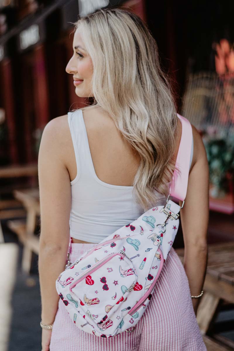 A woman with long blonde hair wears a white tank top and pink striped pants, carrying the Katydid Music Girl Icons Sling Bag with an adjustable strap. She stands outdoors beside wooden benches.