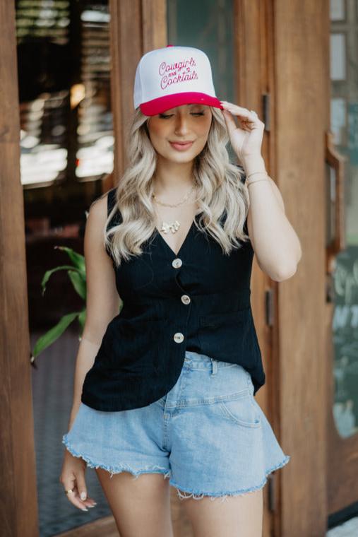 A woman with long blonde hair wearing a red and white cap, black sleeveless top, and denim shorts stands in front of a wooden door with a relaxed expression.