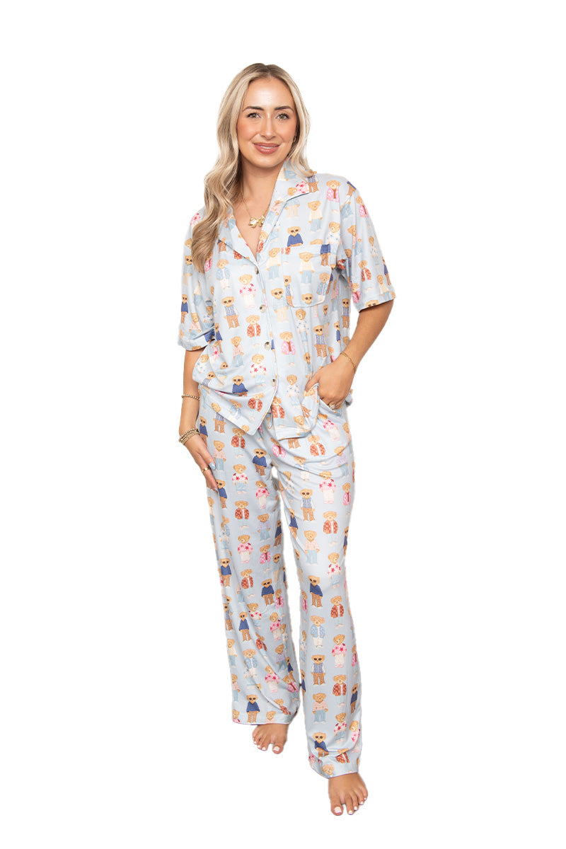 A woman with long blonde hair stands barefoot, smiling at the camera in the Katydid Teddy Bears Womens Cute PJ Set—light blue button-up top and matching pajama pants with a playful print—showcasing cozy loungewear against a white background.