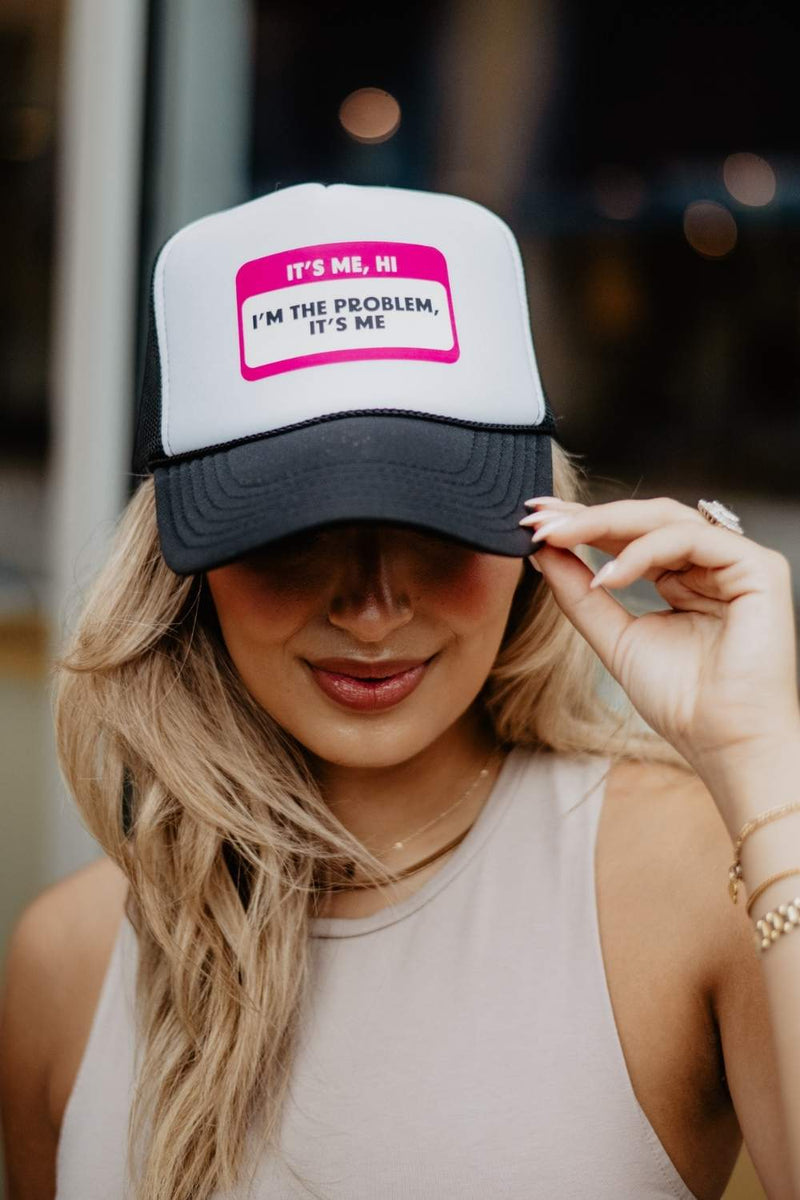 A woman with long blonde hair smiles and gently holds the brim of a Katydid Its Me Hi Im the Problem Its Me Name Tag Hat, featuring black and white colors with pink and black text.