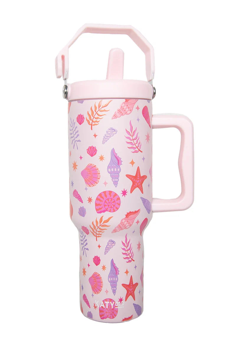The Katydid Beach Seashells Stainless Steel Tumbler is a tall pastel pink travel cup with a large handle, flip-up straw lid, and features colorful seashells, starfish, and seaweed in pink, purple, and orange hues.