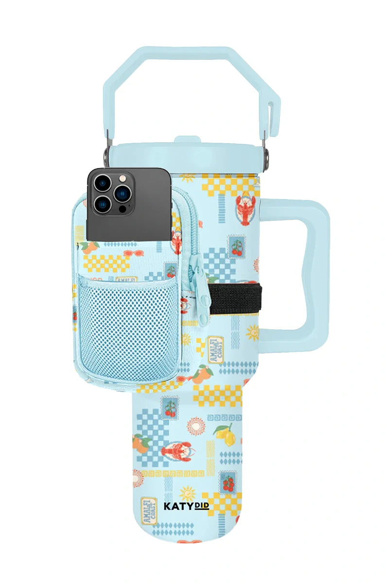 The Katydid Italian Summer Tumbler Belt Bag features light blue color, vibrant patterns, a handle and strap, zippered phone pocket, front mesh pouch, and branding at the bottom—ideal for carrying drinks hands-free.