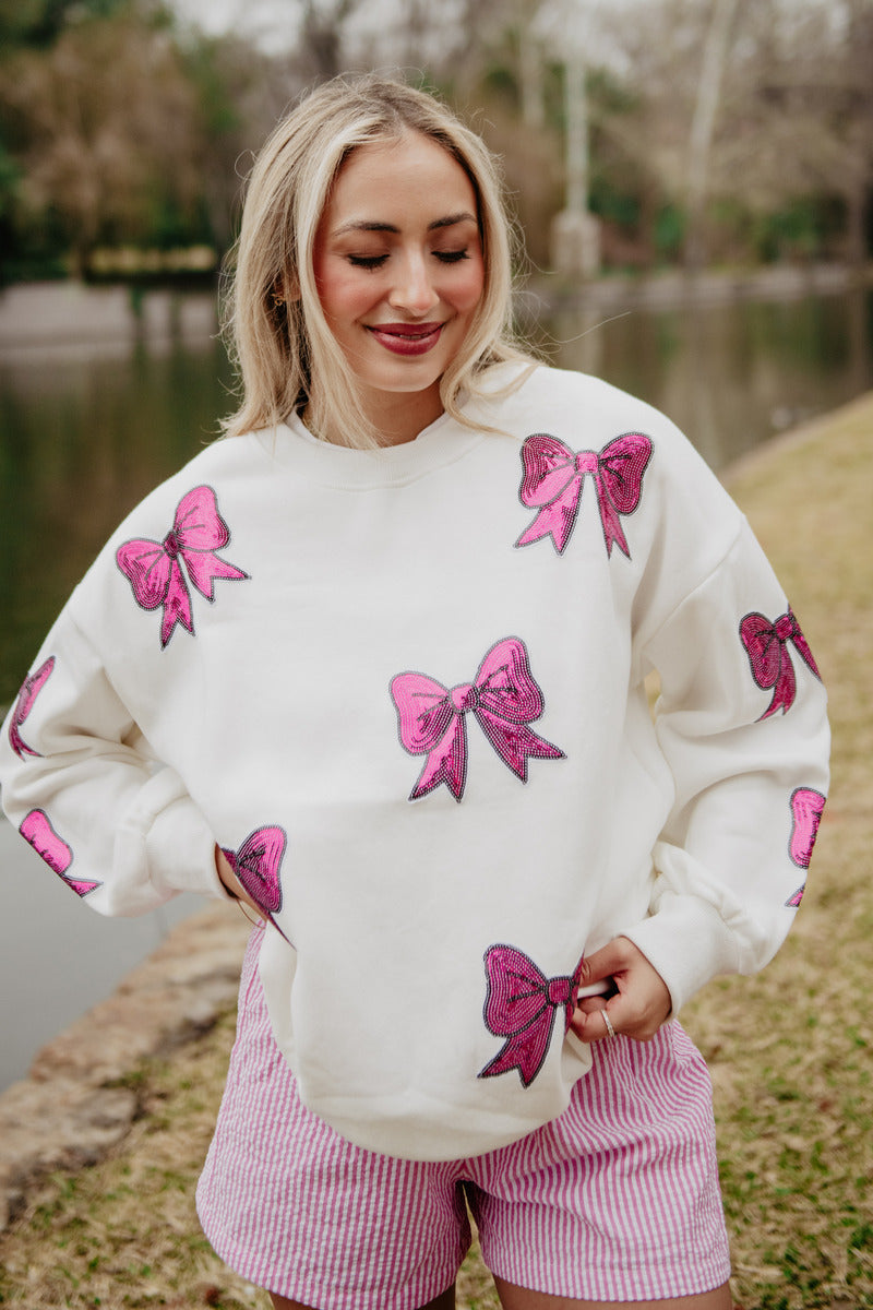 A woman stands outdoors by a canal, smiling with her eyes closed. She wears the Katydid Sequined Coquette BOW Patches Sweatshirt with oversized fit, paired with pink-and-white striped shorts. Grass, trees, and water fill the background.