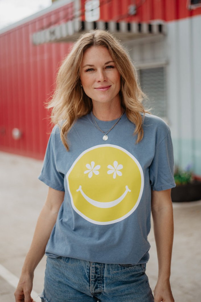 A woman with blonde hair stands outside near a red building, smiling at the camera in the Katydid Daisy Eyes Happy Face Graphic Tee paired with denim shorts.
