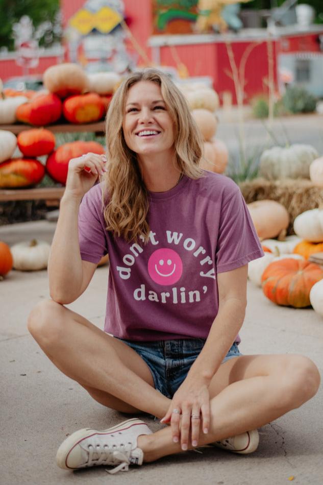 A smiling woman sits cross-legged outdoors by pumpkins, wearing the Katydid Dont Worry Darling Happy Face T-Shirt in purple and denim shorts—one of the popular Comfort Colors tees.