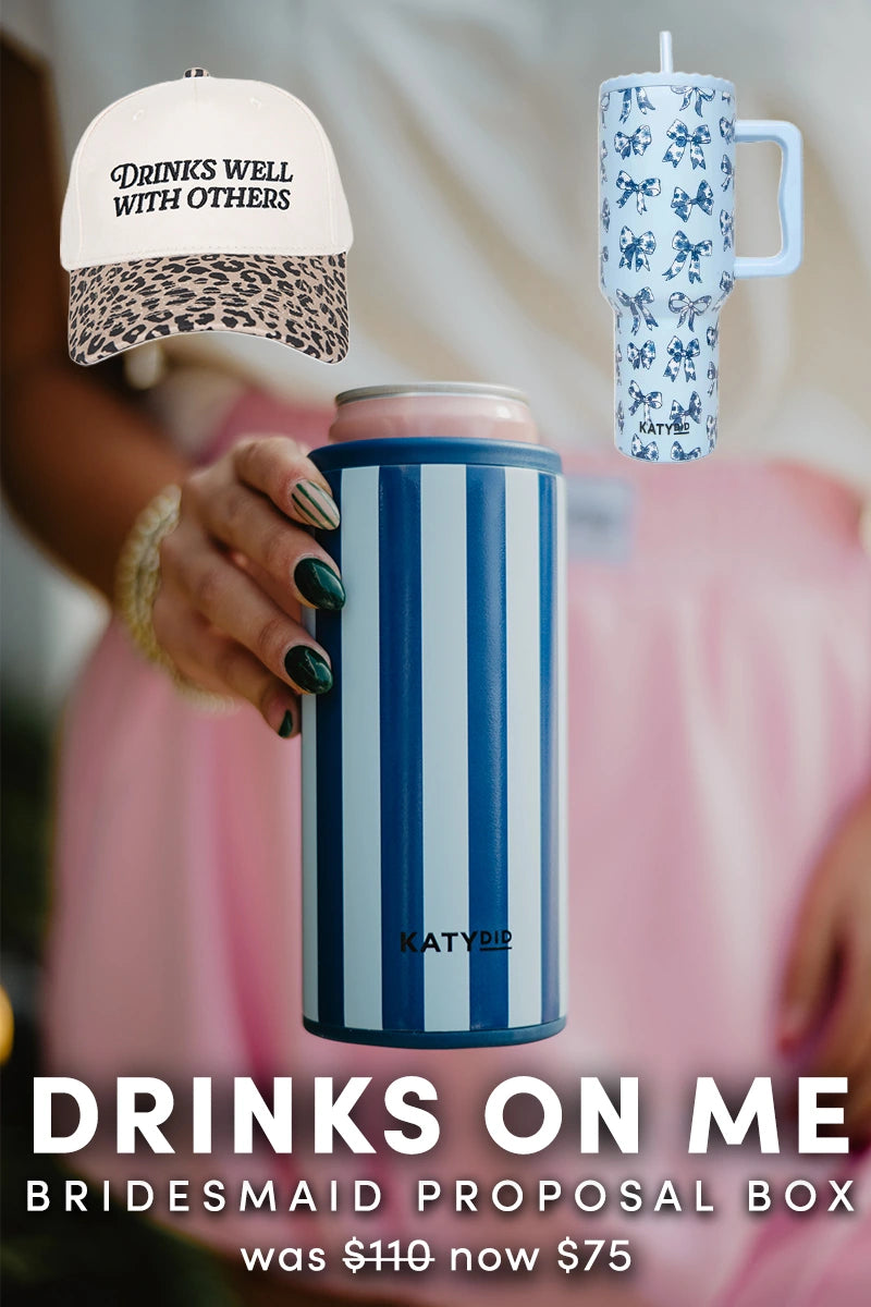 A person holds a blue-striped can cooler. Above are a leopard-print “DRINKS WELL WITH OTHERS” cap and a blue floral tumbler. This Katydid.com Drinks On Me Bridesmaid Proposal Box was 110, now 75—perfect for bridal party gifting.