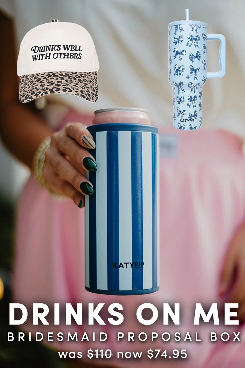 A hand holds a blue-and-white striped can cooler. Above are a cheetah print DRINKS WELL WITH OTHERS cap and blue floral tumbler. Text below promotes the Katydid.com Drinks On Me | Bridesmaid Proposal Box, now on sale for 74.95.