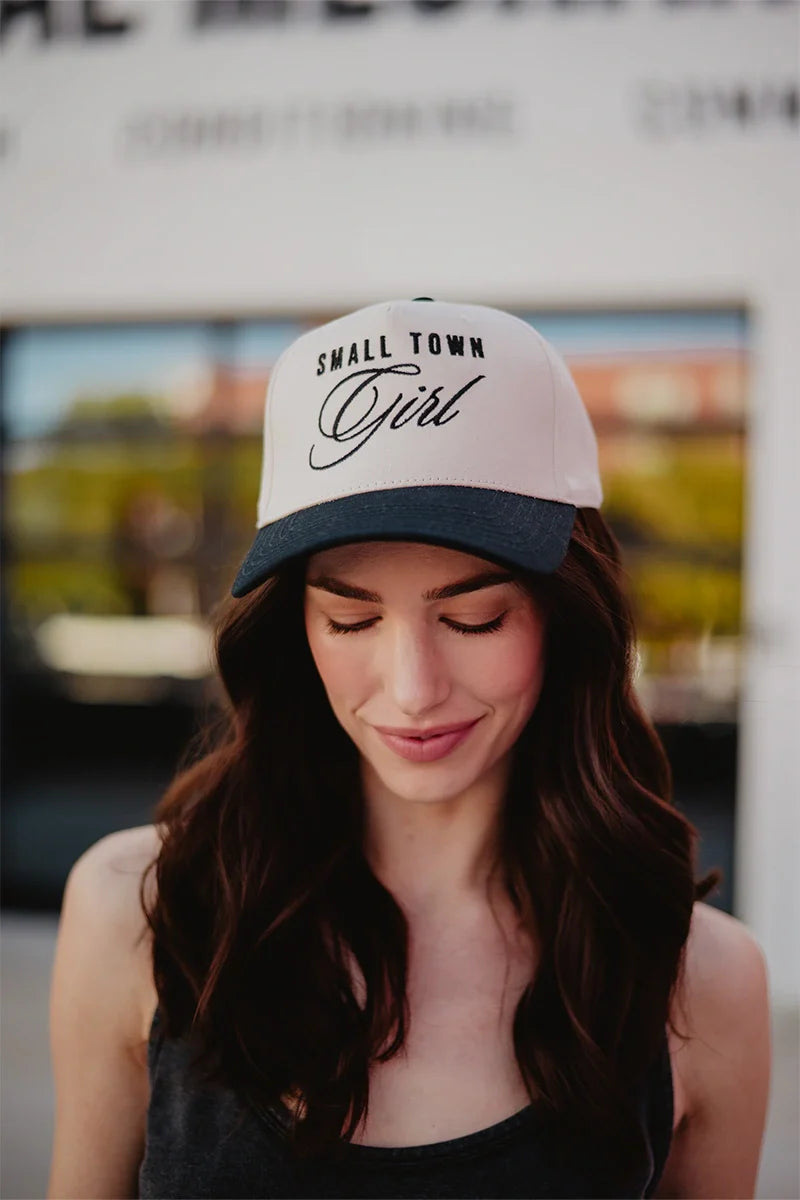 A young woman with long brown hair, wearing a black tank top and a Katydid Small Town Girl Vintage Trucker Hat, stands outdoors with her eyes closed, softly smiling in her country-cute hat.