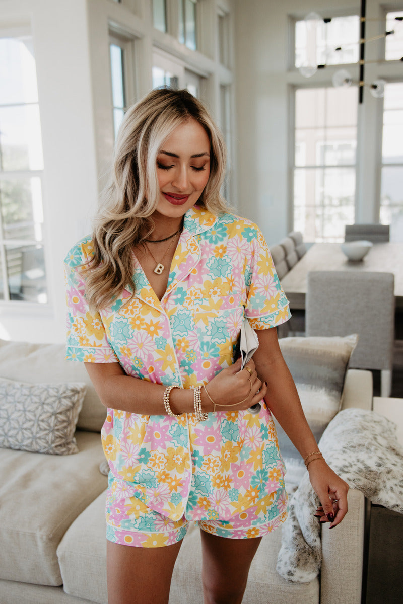 A woman with long blonde hair smiles while wearing colorful floral pajamas in a bright, modern living room, conveying a relaxed and cheerful mood.