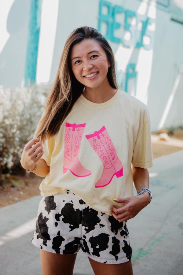 A woman with long brown hair smiles outdoors on a sunlit sidewalk, wearing the Katydid Kicking Boots Western Cowgirl T-Shirt featuring pink cowboy boots and black-and-white cow print shorts.
