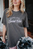 A woman with wavy blonde hair stands outdoors in front of a brick building, holding black and silver pom-poms and wearing the Katydid Game Day Social Club Graphic Tee.
