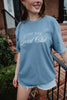 A woman stands outside on brick steps, smiling and wearing the Katydid Game Day Social Club Graphic Tee in blue with white script, paired with light denim shorts.