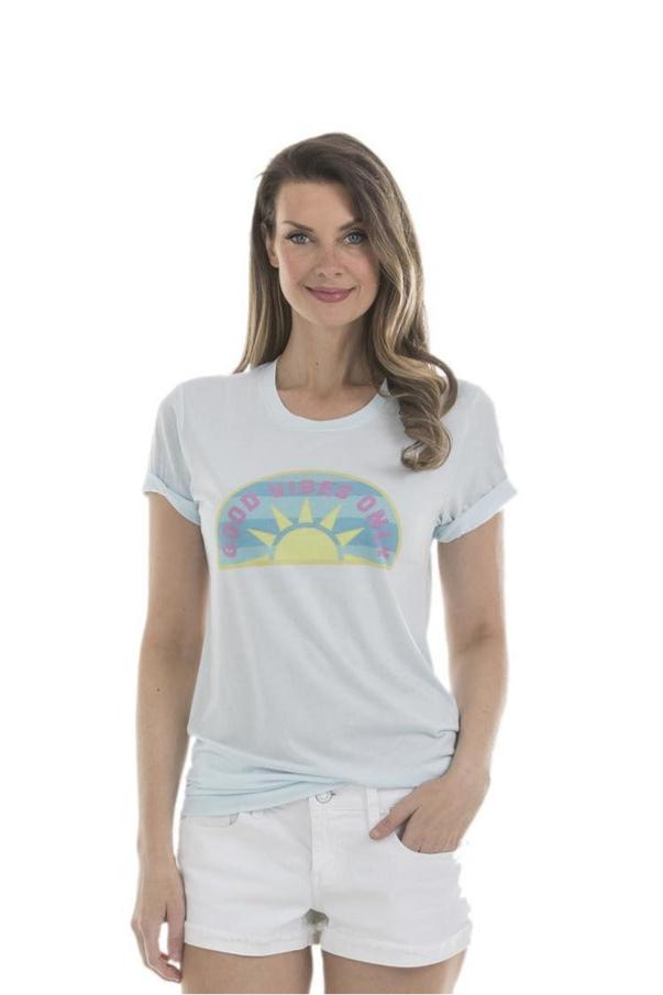 A smiling woman with long brown hair models the Katydid Good Vibes Only T-Shirt, featuring a light blue round neck, yellow sun graphic, and bold text, paired with white shorts against a white background.