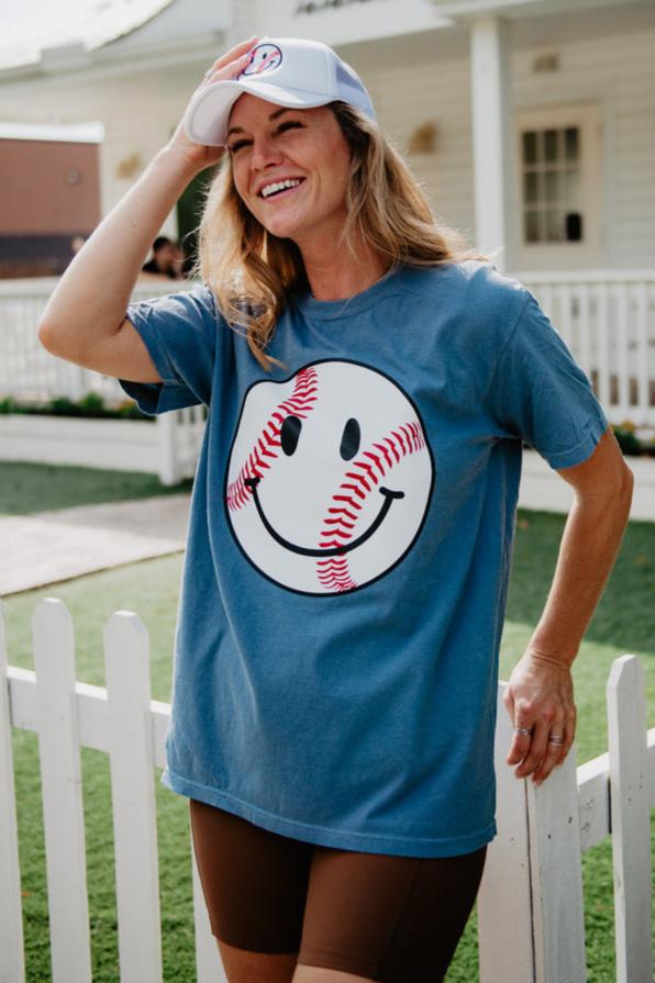A woman smiles by a white picket fence, wearing Katydids Baseball Happy Face Cute Graphic Tee, a white cap, and brown shorts. A house is visible in the background.