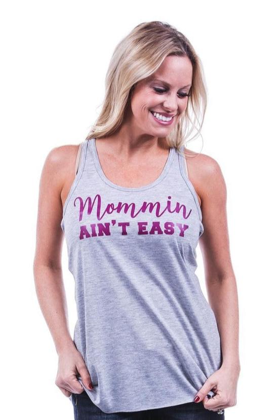 A smiling woman with long blonde hair wears a Katydid Mommin Aint Easy tank, featuring purple and pink lettering, standing against a plain white background.