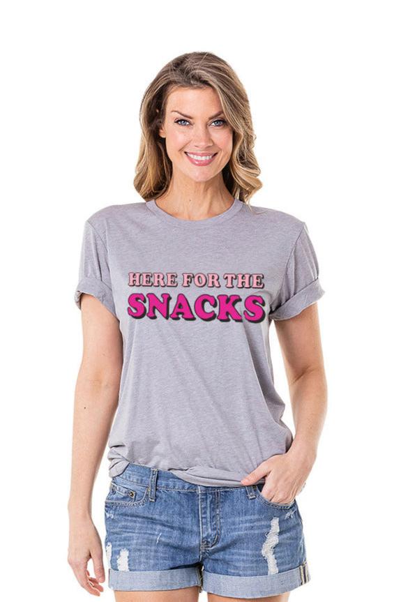 A woman wears the Katydid Here For The Snacks Graphic Print T-Shirt, with “HERE FOR THE SNACKS” in pink and red on light gray, paired with denim shorts as she smiles against a plain white background.