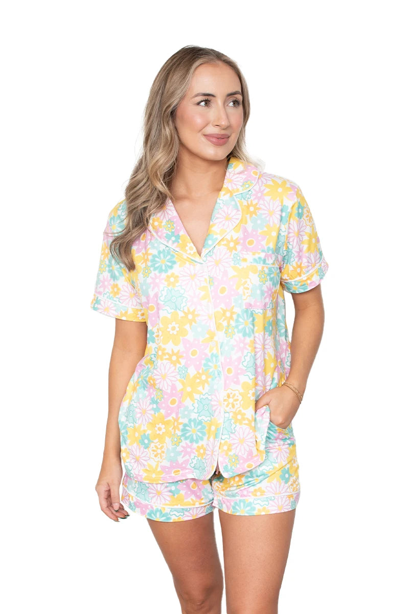 A woman wears the Katydid Spring Flowers Pajama Shorts Set, featuring a colorful yellow, pink, blue, and white floral pattern. This button-up top and matching shorts loungewear makes a perfect Mother’s Day gift.