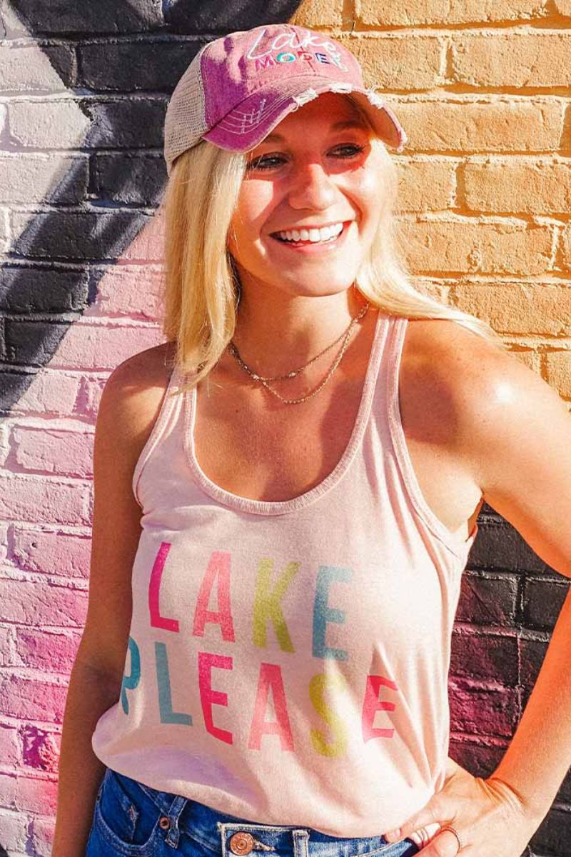 A smiling woman stands before a colorful brick wall, wearing Katydid’s Lake Please Womens Tank Top in light pink with bold multicolored text and a matching cap. Sunlight highlights her blonde hair.