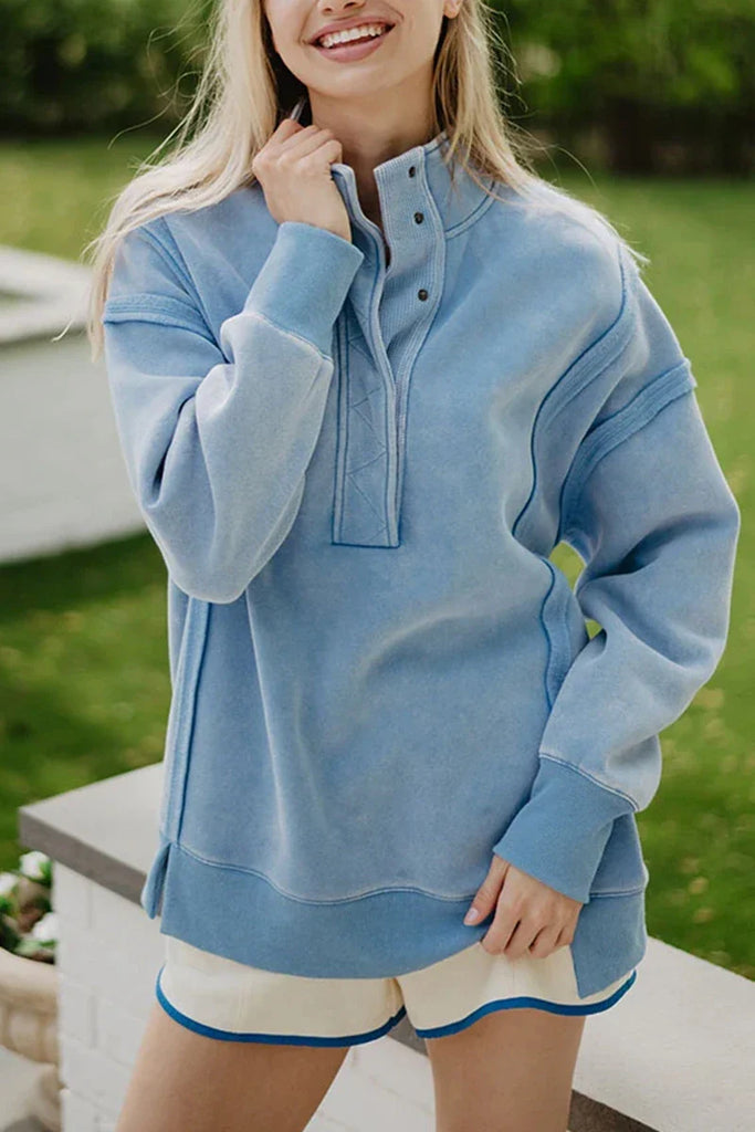 Outdoors, a person smiles with one hand on a bench, wearing the Katydid Light Blue Vintage Wash Snap-Neck Pullover and cream shorts with blue trim.