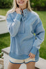 Outdoors, a person smiles with one hand on a bench, wearing the Katydid Light Blue Vintage Wash Snap-Neck Pullover and cream shorts with blue trim.