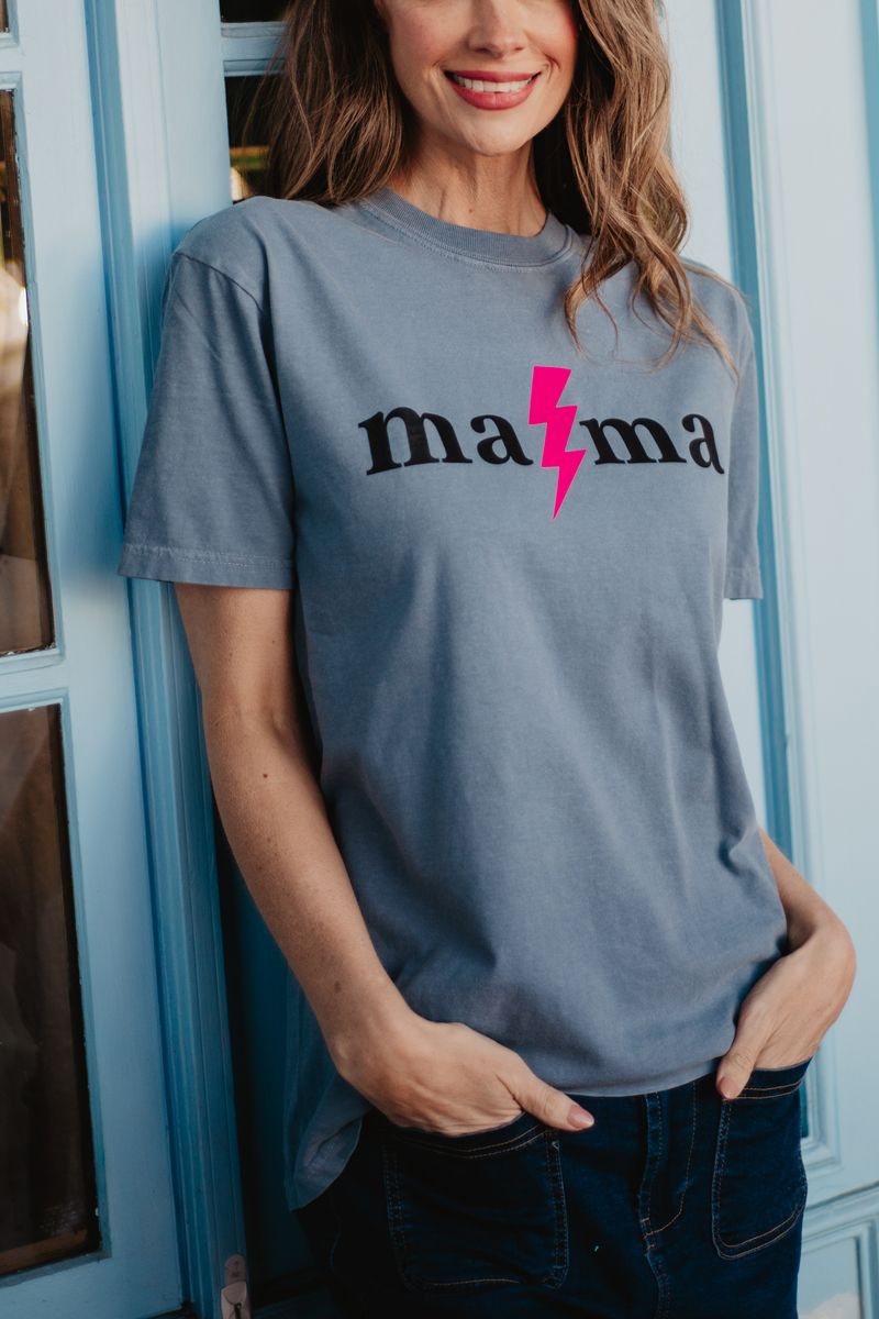 A woman in a gray tee with “mama” in black and a pink lightning bolt on the second “m” stands smiling by a blue wall—an ideal Mother’s Day gift from Katydid.com, especially when included with the Moms Faves Gift Box.