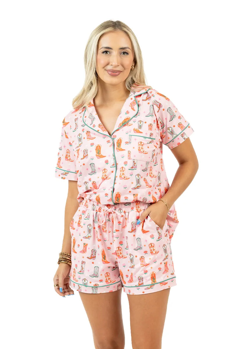 A woman with long blonde hair smiles against a white background, wearing Katydid’s Boots  Roses Pajama Set—a pink, short-sleeve button-up shirt and shorts adorned with colorful cat illustrations for comfy western-style lounging.