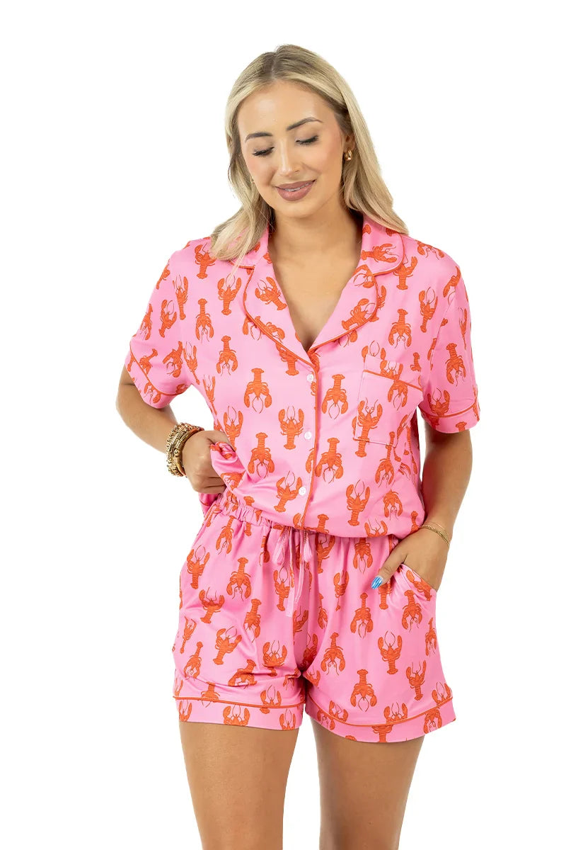 A woman wearing Katydid’s Lobsters Womens Pajama Shorts featuring a pink and orange lobster print smiles with her eyes closed, one hand in her pocket, against a white background.