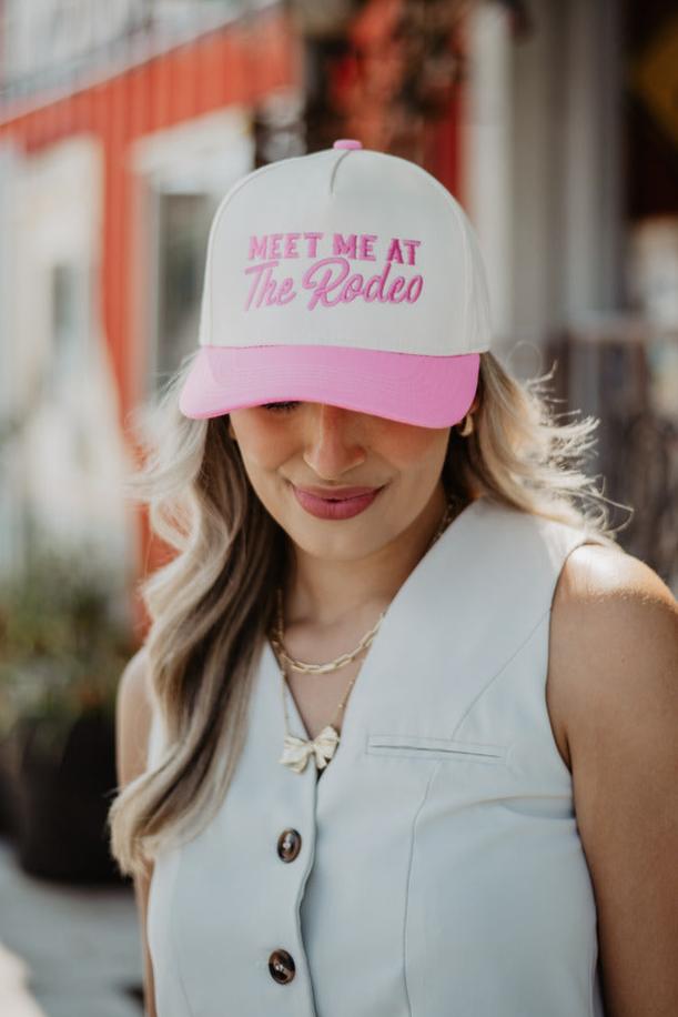 A woman in a light-colored vest smiles, wearing a pink and white cap that reads "Meet Me at The Rodeo." The background features a blurred red and white building.