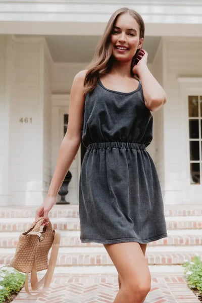 A woman in the Katydid Black Summer Dress stands outdoors on brick steps, smiling and holding a woven handbag in front of a white house with large windows and lush greenery.