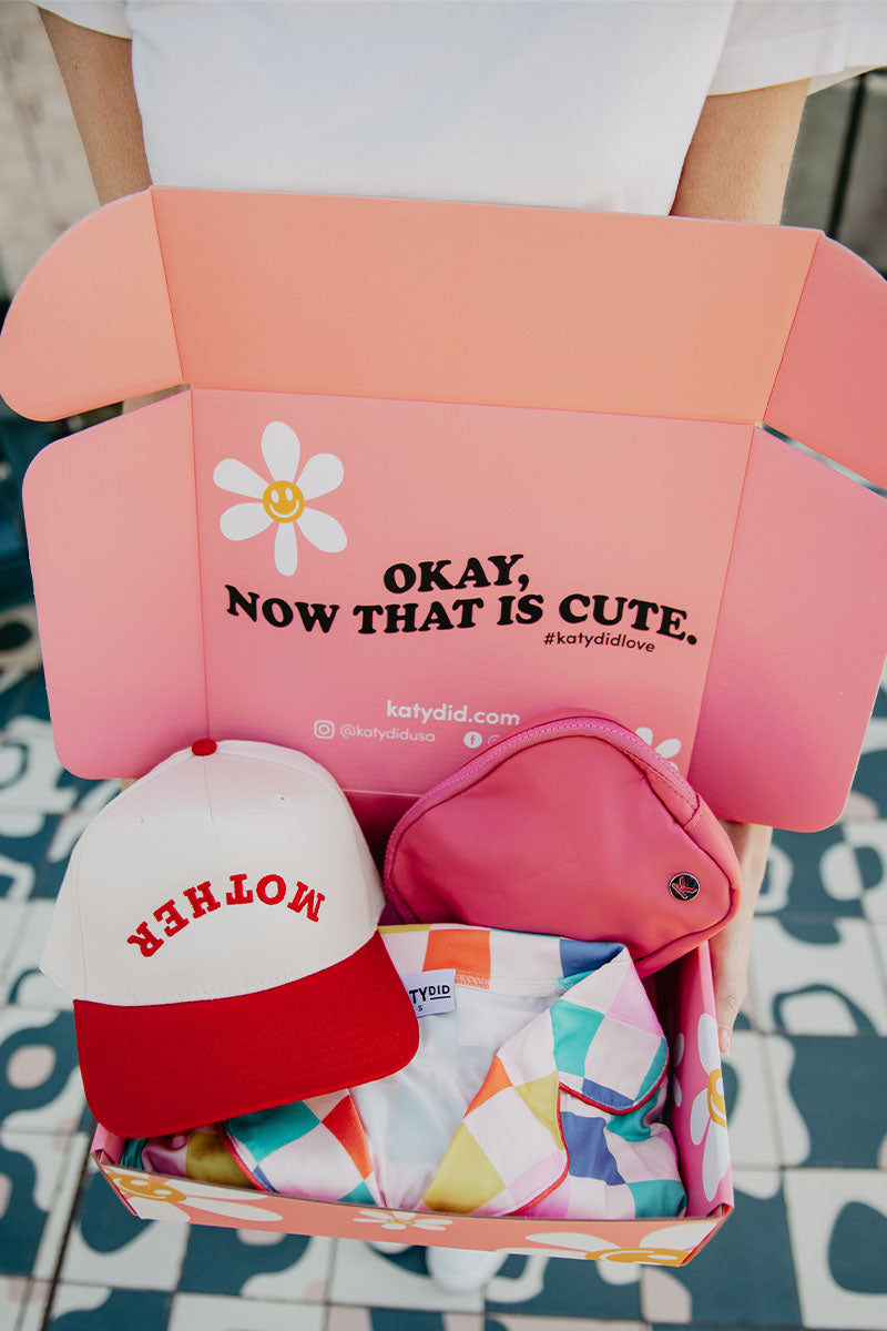 A person holds the Made for Mom Gift Box from Katydid.com, open to reveal a white and red “MOTHER” cap, pink bag, and patterned shirt—an ideal Mothers Day gift set.
