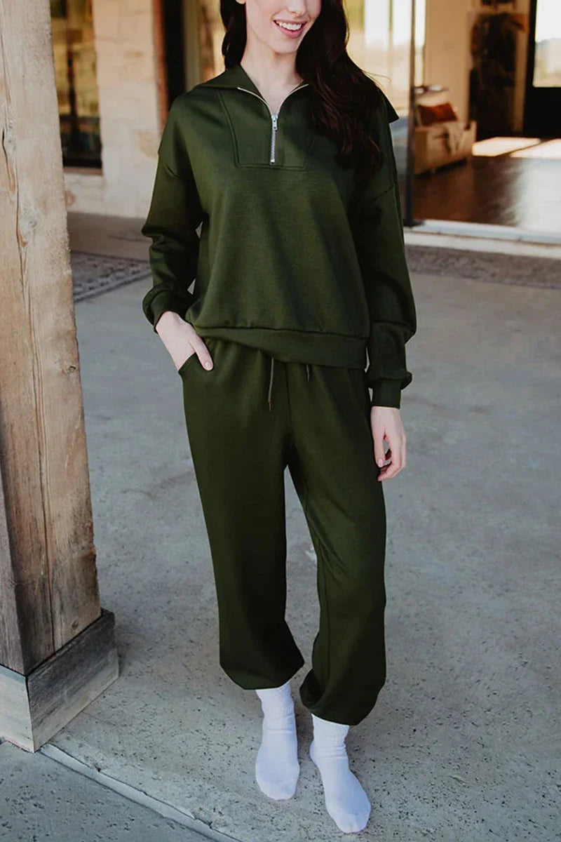 A woman stands on a concrete porch in the Katydid Olive Pullover Two-Piece Set—an olive green zip-up sweatshirt and matching jogger pants. She has her hands in her pockets, wearing white socks, with long brown hair, her face partially out of frame.