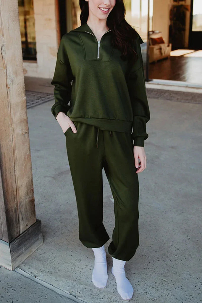 A woman stands on a concrete porch in the Katydid Olive Pullover Two-Piece Set—an olive green zip-up sweatshirt and matching jogger pants. She has her hands in her pockets, wearing white socks, with long brown hair, her face partially out of frame.