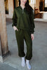 A woman stands on a concrete porch in the Katydid Olive Pullover Two-Piece Set—an olive green zip-up sweatshirt and matching jogger pants. She has her hands in her pockets, wearing white socks, with long brown hair, her face partially out of frame.