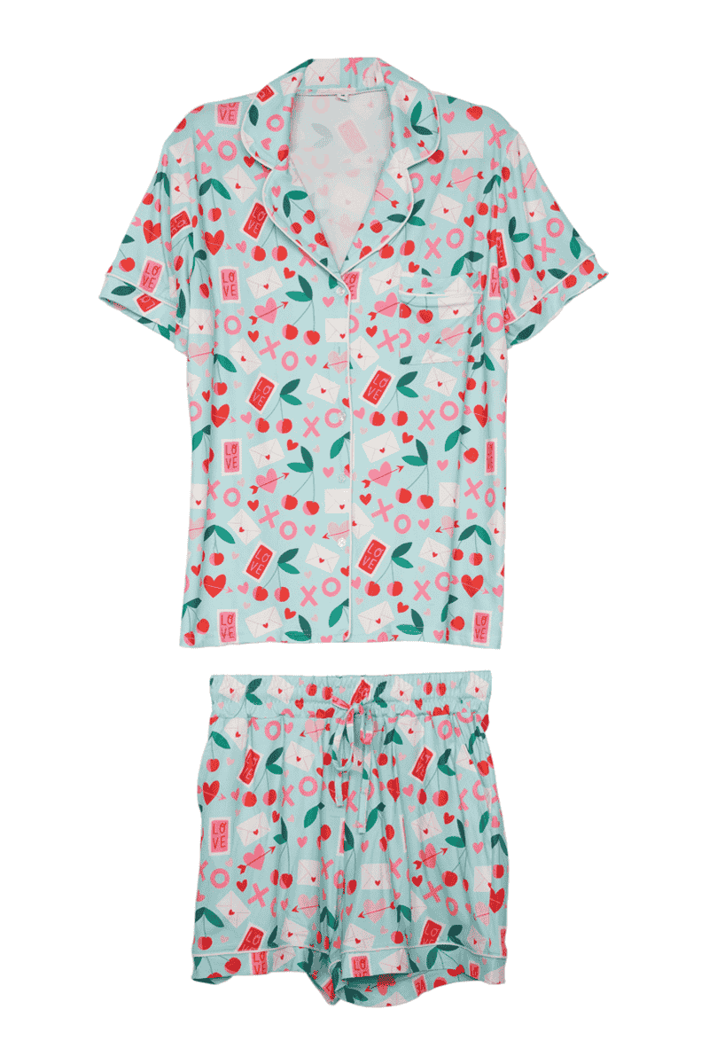 The Katydid XO Cherries Valentines Day Pajamas feature a short-sleeve button-up top and matching shorts with a light blue background and pink, red, and green heart and love-themed prints—perfect for Valentine’s Day.