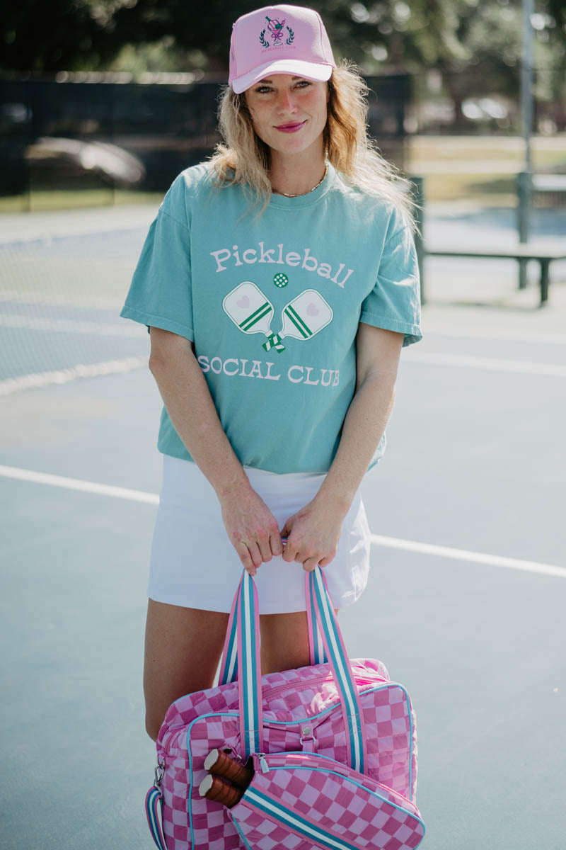 Woman on a tennis court holding a pink checkered bag. She wears a teal "Pickleball Social Club" shirt, white skirt, and pink cap, smiling casually.