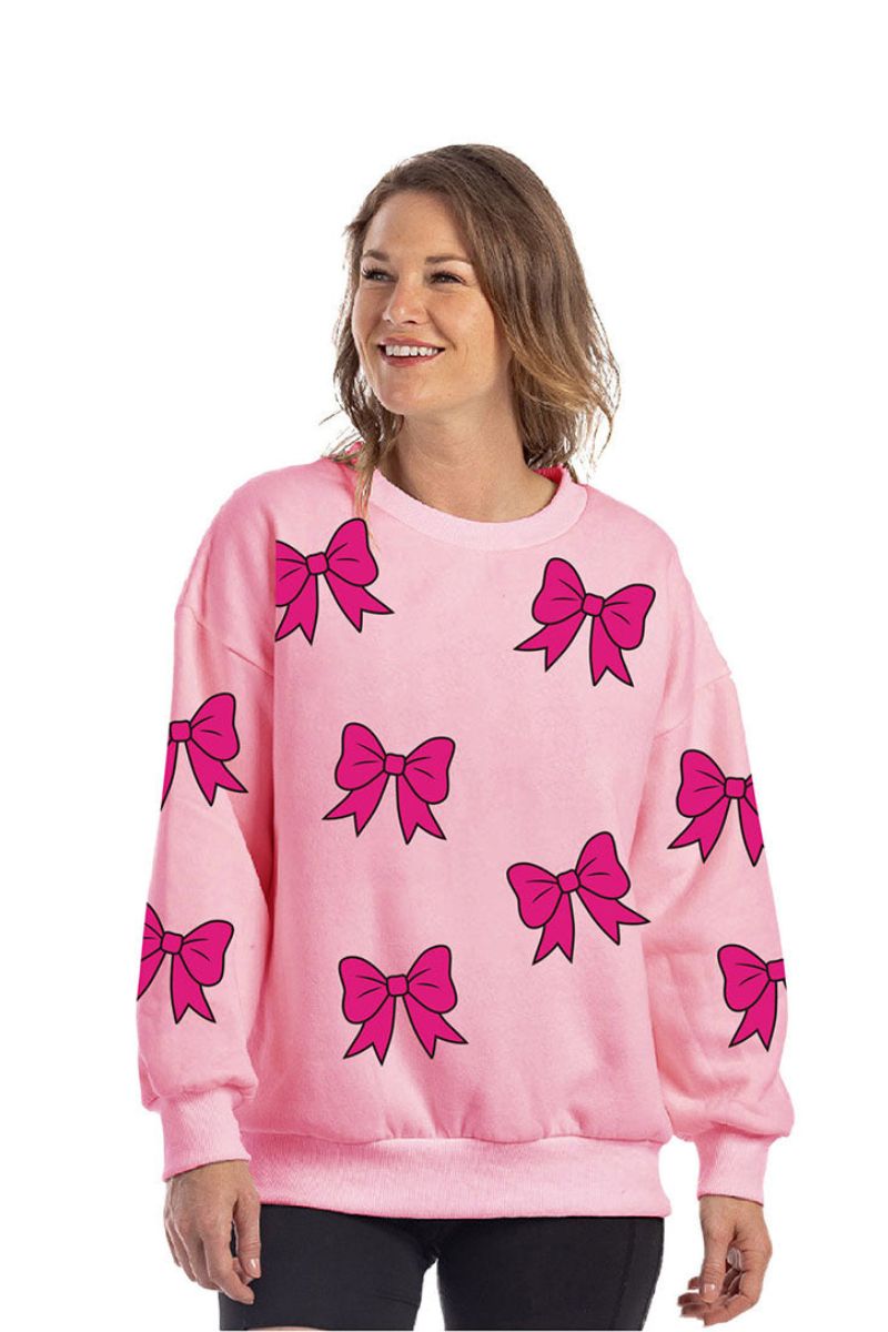 A woman with light brown, wavy hair smiles in a Katydid Sequined Coquette BOW Patches Sweatshirt—an oversized, light pink top adorned with large sequined bow designs—while standing against a plain white background.