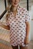 A woman with wavy blonde hair stands indoors, holding a stair railing. She wears the Katydid Black  Pink Mini Coquette Bows Loungewear Set, with part of a staircase visible behind her.