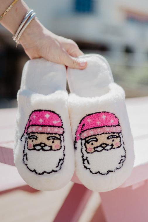 A hand holds fluffy white slippers embroidered with Santa's face wearing a pink hat.