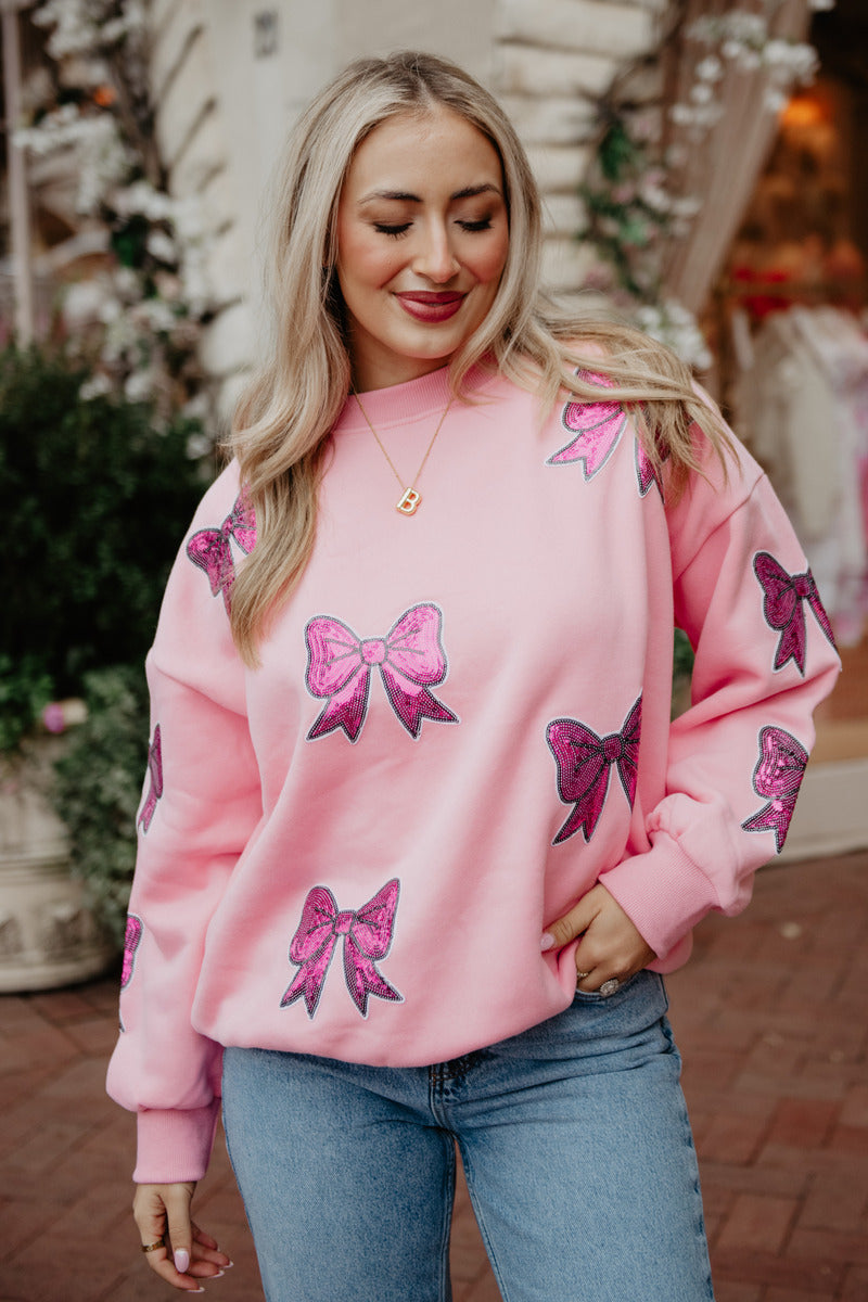 A woman with long blonde hair wears the Katydid Sequined Coquette BOW Patches Sweatshirt, featuring glittery pink bows, paired with light blue jeans. She smiles outdoors near white flowers and greenery, one hand on her hip.