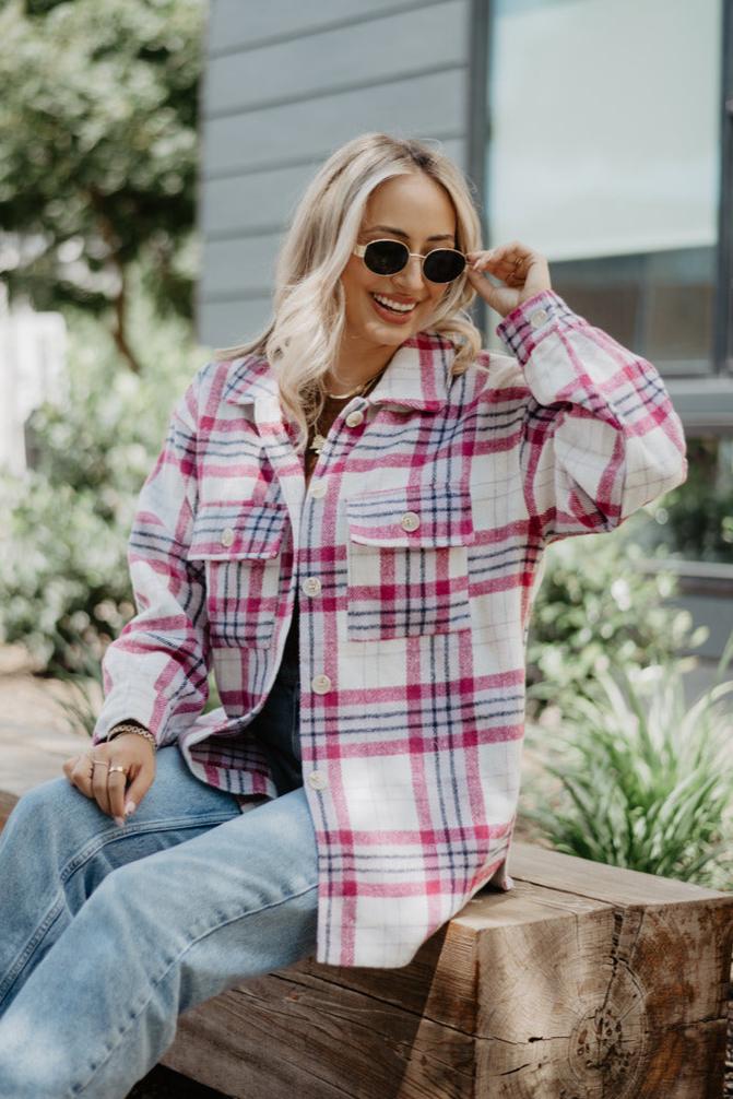 A woman with long blonde hair and sunglasses smiles outdoors, wearing the Katydid Magenta/Purple Plaid Shacket for Women as she sits on a wooden bench amid greenery and a modern building.