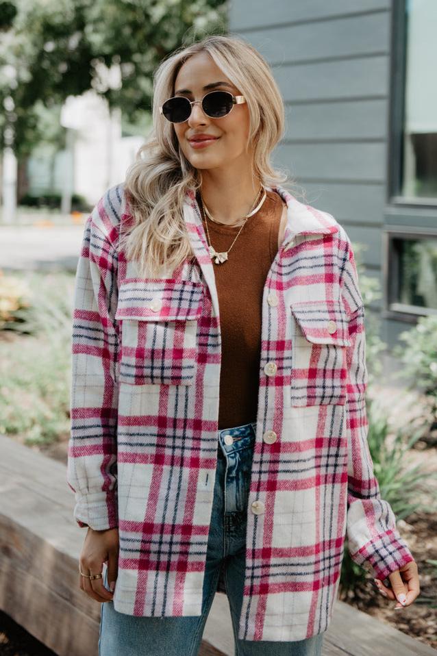 A woman with long blonde hair wears sunglasses, a brown top, blue jeans, and the Katydid Magenta/Purple Plaid Shacket for Women, standing outdoors in front of greenery and a modern building.
