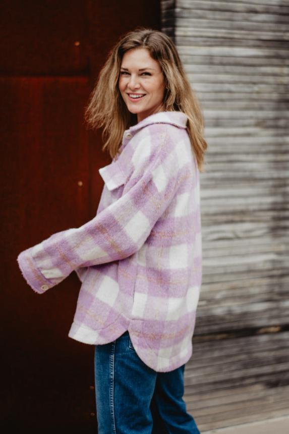A woman with wavy hair, wearing the Katydid Purple Plaid Checker Shacket and blue jeans, smiles as she looks over her shoulder outdoors against a wooden wall and a brown metal surface.