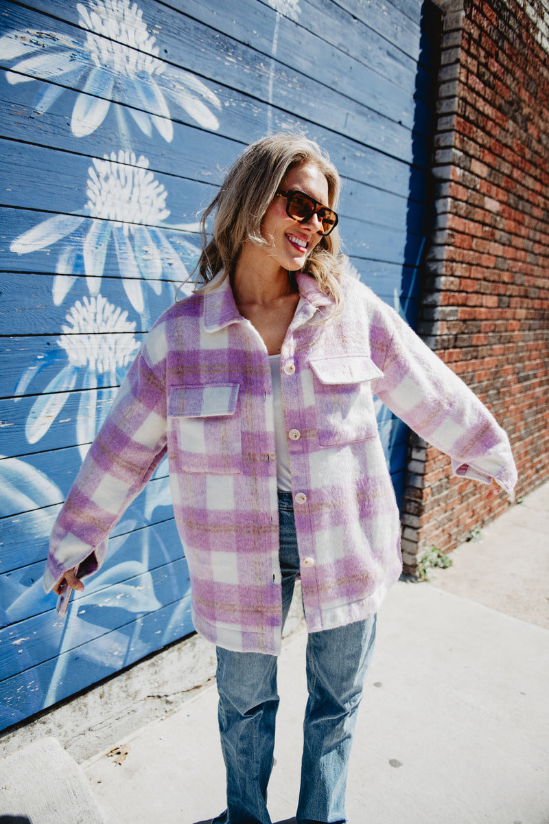 A person in the Katydid Purple Plaid Checker Shacket and blue jeans smiles with arms outstretched in front of a blue mural with large white flowers and a brick wall, wearing sunglasses.