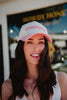 A smiling woman with long dark hair wears a white and pink PLEASE BE PATIENT WITH ME, IM FROM THE POOL cap—an ideal Mother’s Day gift or cozy addition to The Tired Mama Gift Box by Katydid.com—while standing outside a storefront.