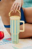 A person in a blue swimsuit by a poolside holds a pastel blue and yellow striped tumbler with a handle. A splash of red, possibly a drink, is visible.