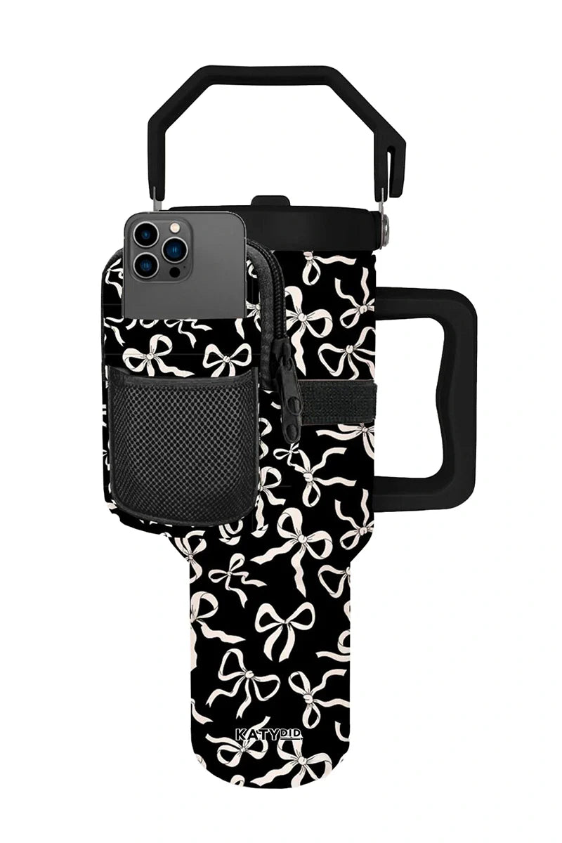 The Katydid Black  White Bows Tumbler Pouch features a handle, front phone pocket, and cute white bow patterns with the brand name at the bottom. A smartphone fits perfectly in the front pocket of this stylish pouch.